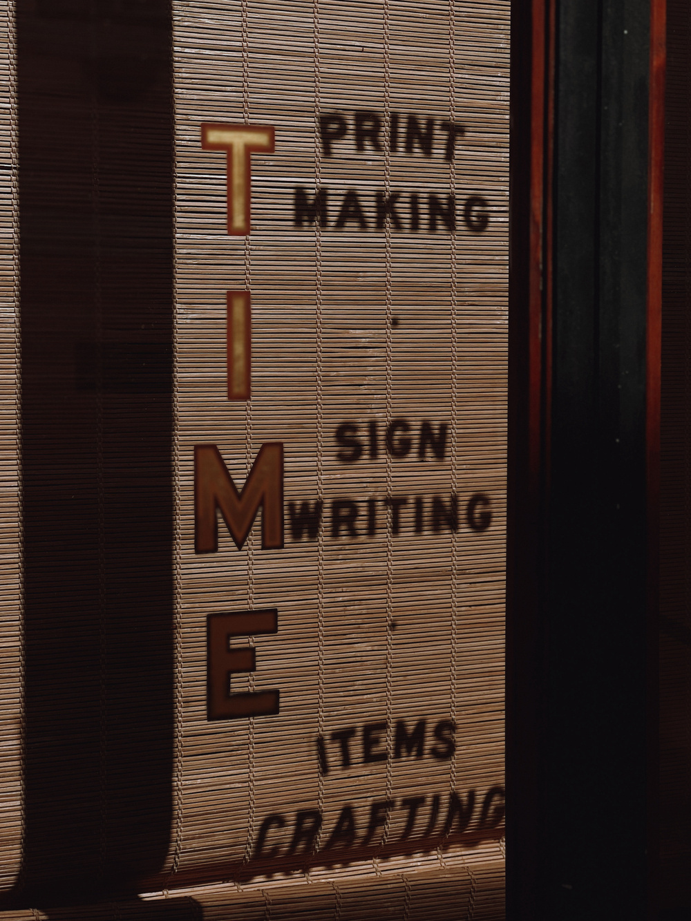 Vertical blinds with sunlight casting shadows. Large, stylized letters spell “TIME,” with smaller text reading “PRINT MAKING,” “SIGN WRITING,” and “ITEMS CRAFTING."