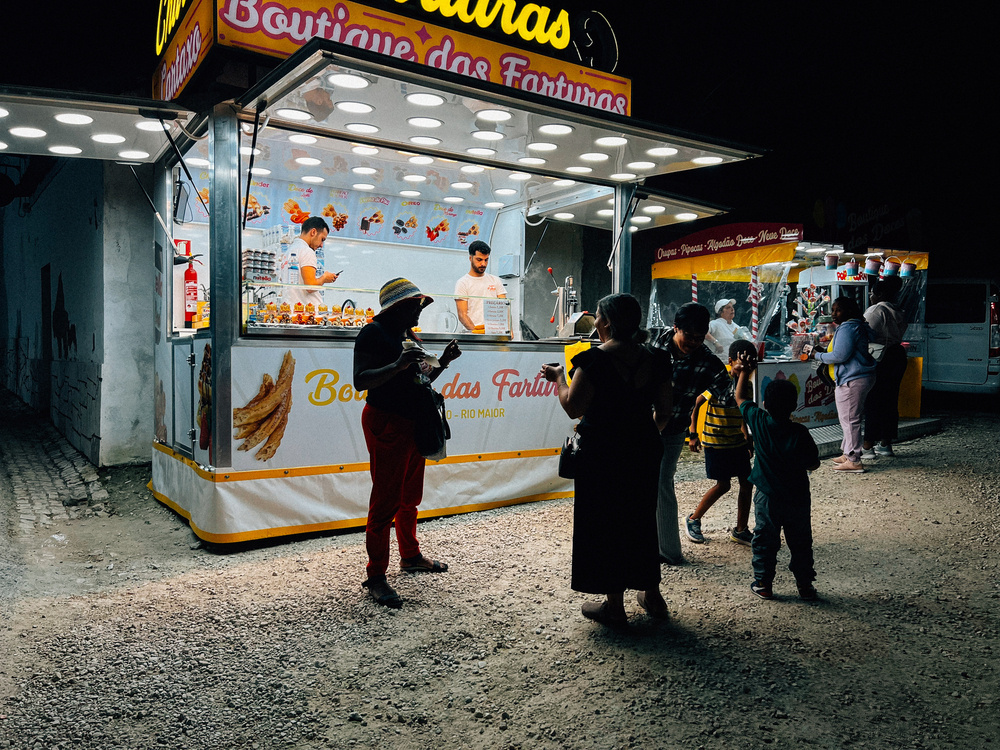 A night scene at a street food kiosk with customers standing in front of it, some are interacting while others are waiting for their orders. Two workers can be seen inside the kiosk. The brightly lit sign reads “Boutique das Farturas”.