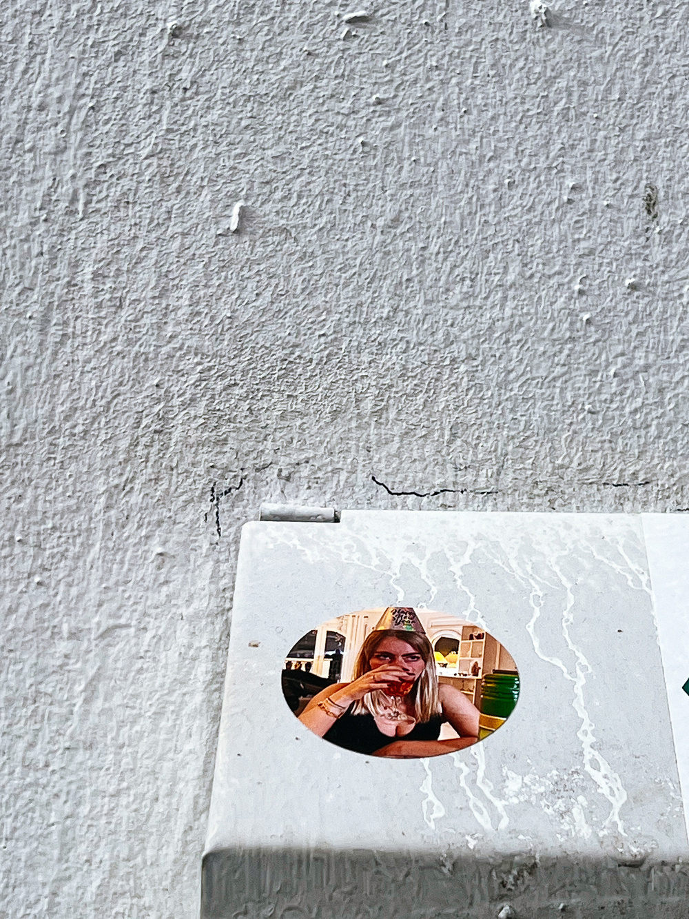 A sticker with a circular photo of a person sipping a drink is affixed on a worn, textured surface.