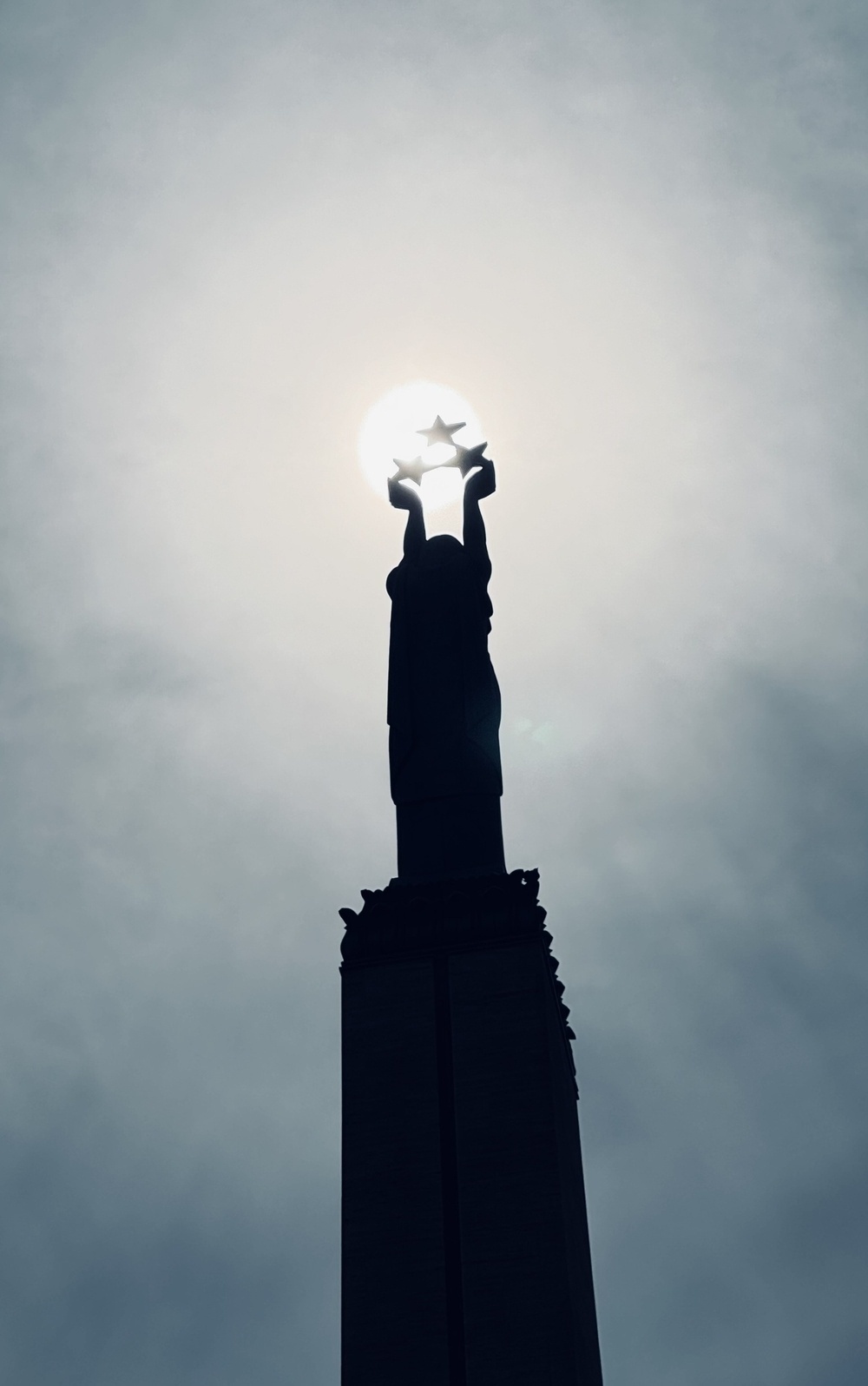 A statue with arms held up to the sky, holding three stars in front of the sun.