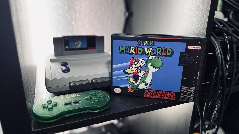 Auto-generated description: A Super Nintendo Entertainment System with a green controller and a boxed Super Mario World game is displayed on a shelf.