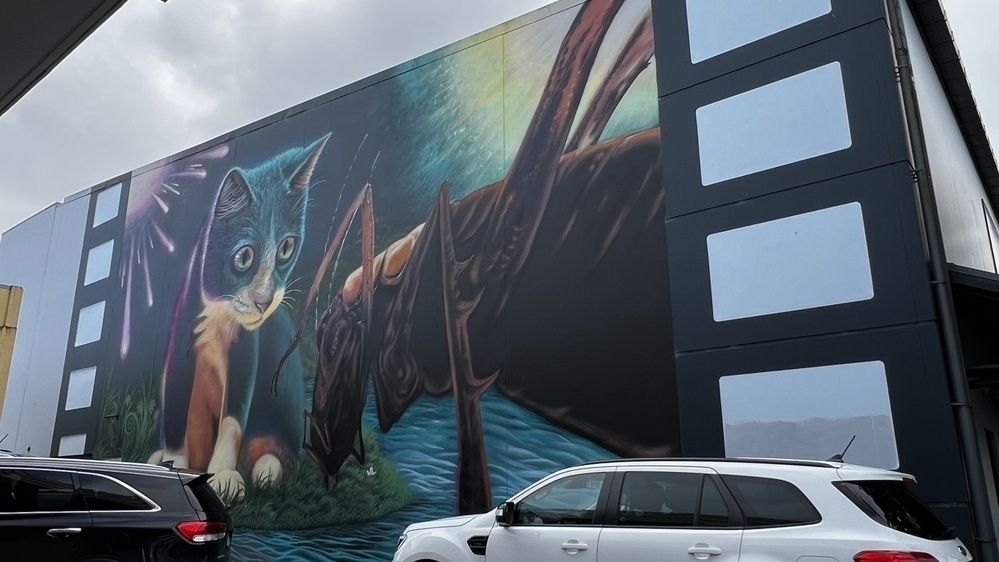 A large mural depicts a surreal scene of a cat with large, round eyes near a towering insect-like creature against a backdrop of an expansive sky, with parked cars in the foreground.