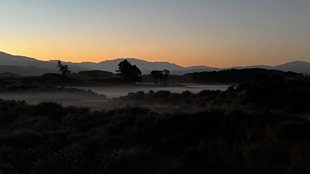 Dark landscape with a puddle of white ground fog and hills behind with a touch of pink in the sky. 