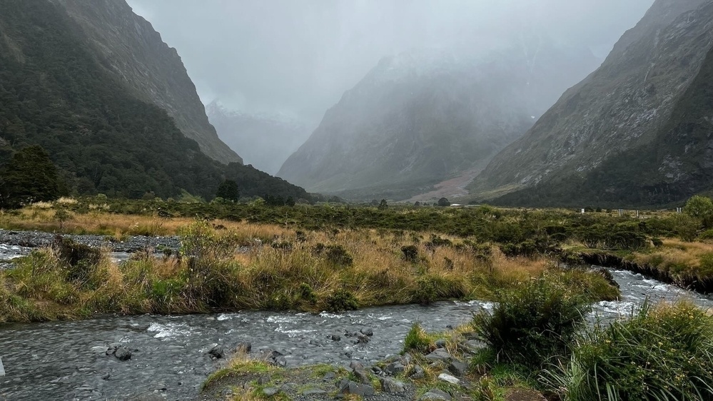 A small creek runs from the foreground towards a misty valley between high mountains. 