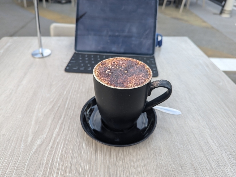 A cappuccino in a black coffee mug, placed on a matching saucer with a spoon beside it, sits on a wooden table outdoors alongside an iPad.
