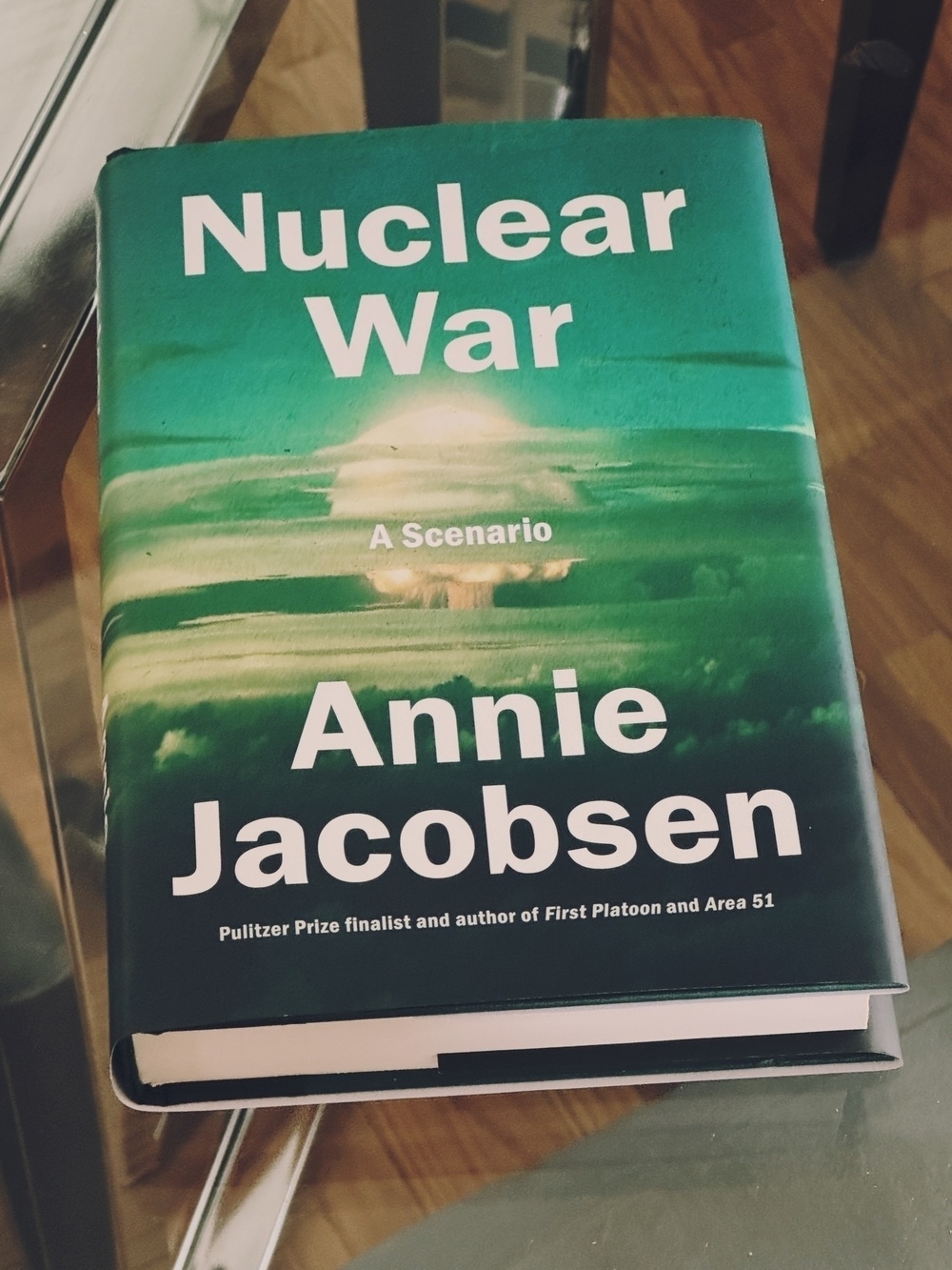 A greenish-black book cover with a nuclear explosion in the background on a glass table - the book is Nuclear War by Annie Jacobsen