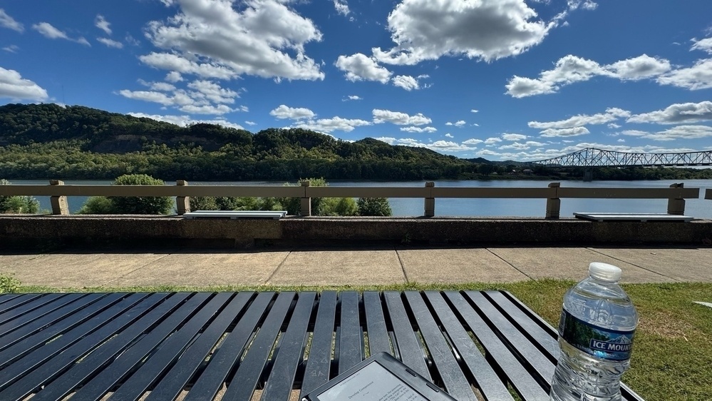 The view of the Ohio River from Alexandria Point Park in Portsmouth, OH. A picnic table with a book and water bottle along with trees, a river and white clouds in a blue sky. 