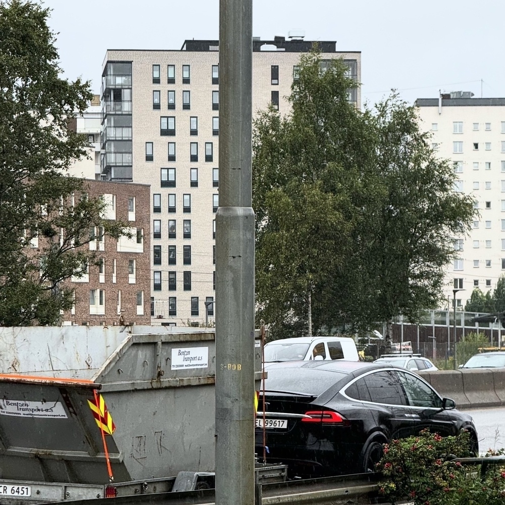 A street features a car (Tesla) towing a Cybertruck, and several residential buildings in the background with trees and a lamppost in the scene.