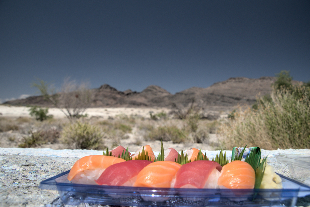 The photo shows an open box of supermarket sushi on a bench in a desert environment.