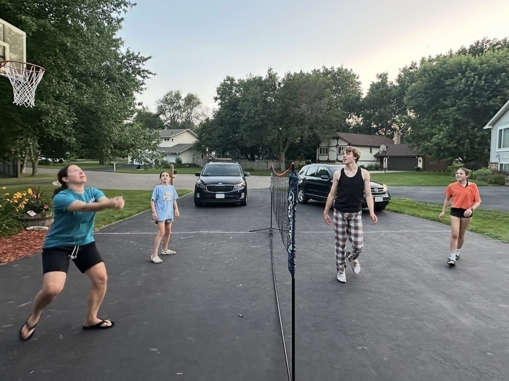 Four people are playing badminton on a driveway with a low net in the center, surrounded by houses, trees, and parked cars.