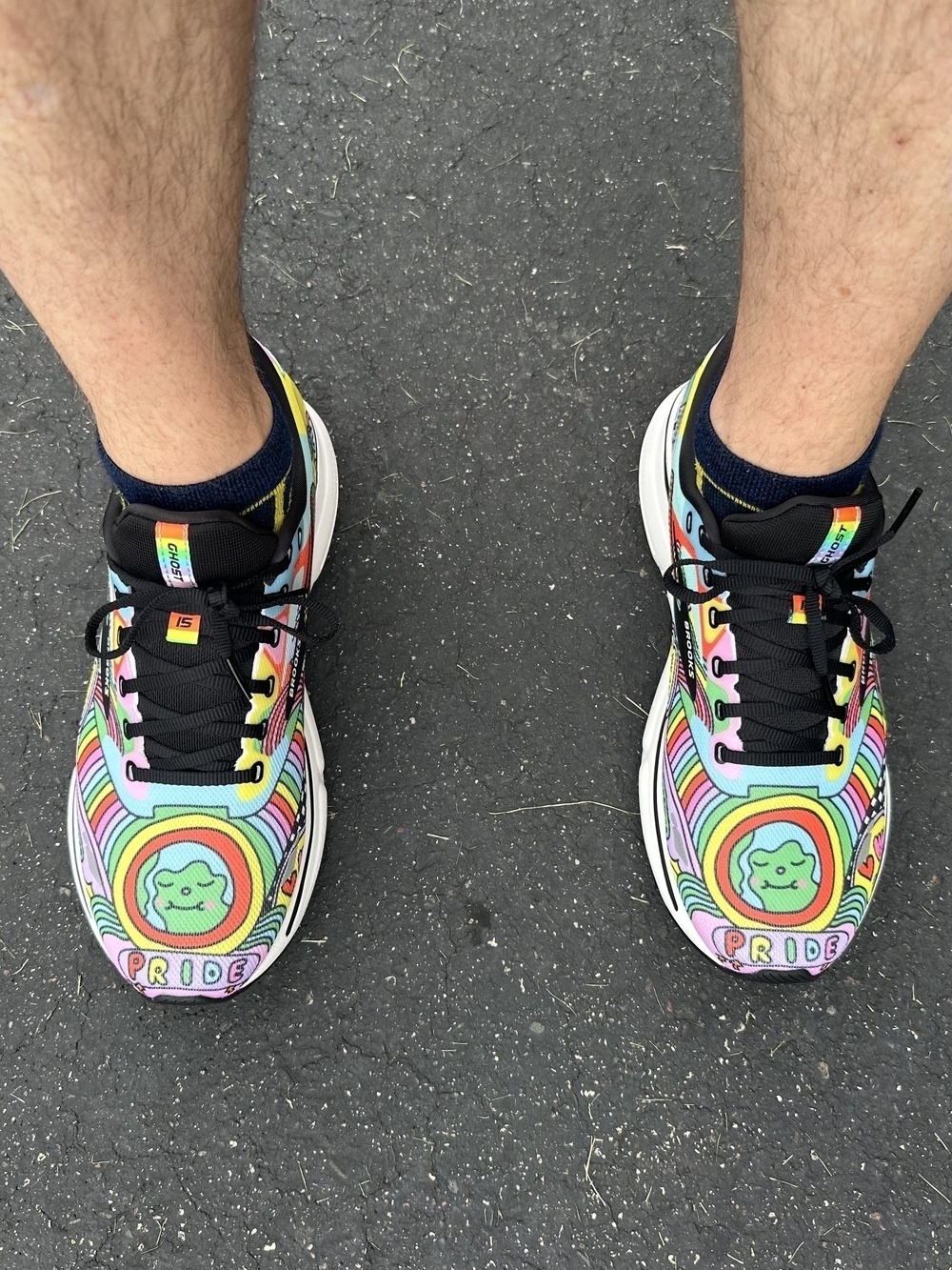 Colorful sneakers featuring rainbow patterns, “PRIDE” text, and a frog graphic; worn by a person with bare legs and black socks, standing on a gray asphalt surface.