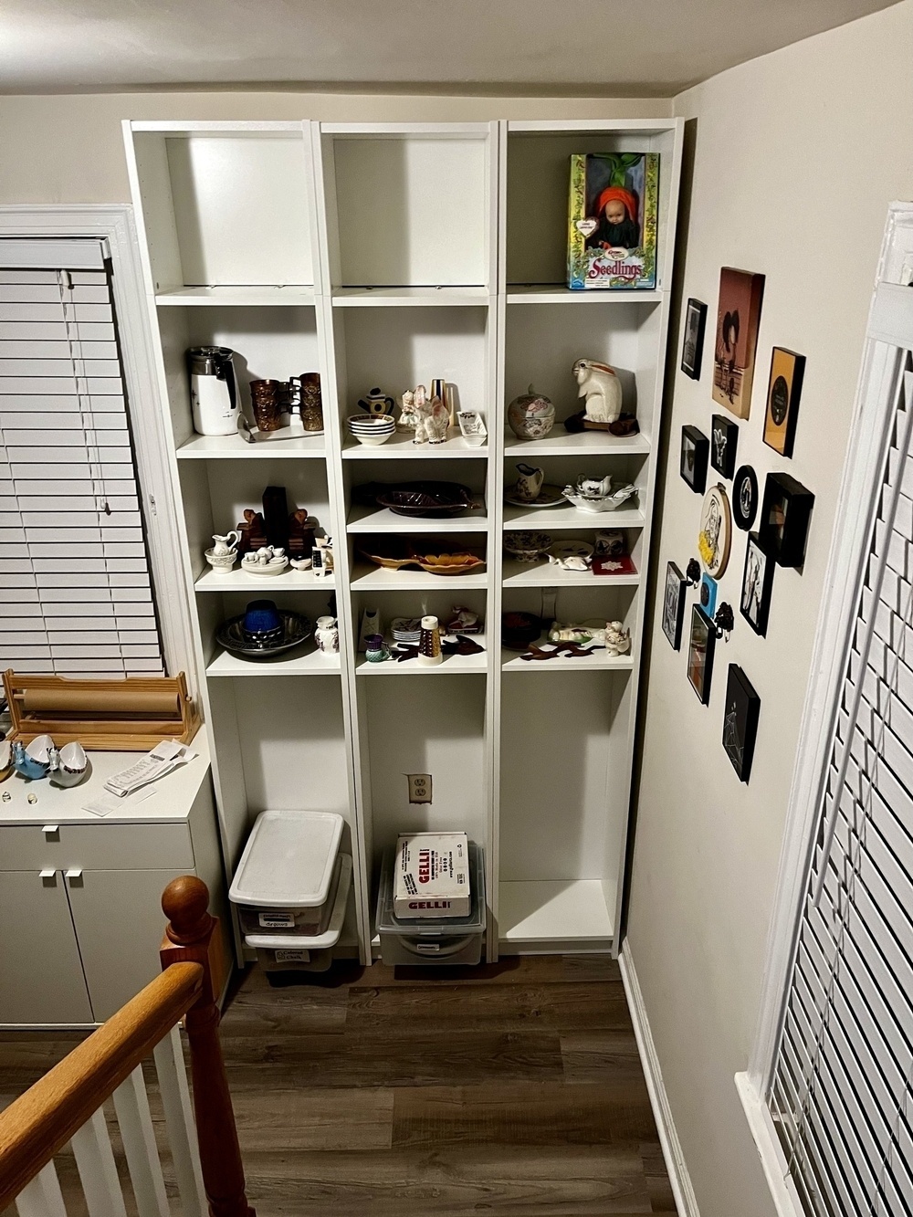 White shelving unit with various items displayed, near a staircase and a window with blinds, in a home interior.