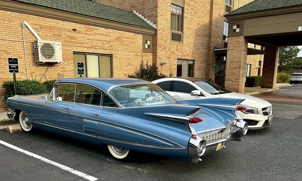 A vintage blue Cadillac with prominent tailfins is parked in a lot beside a modern white car, against a brick building with a covered entryway.