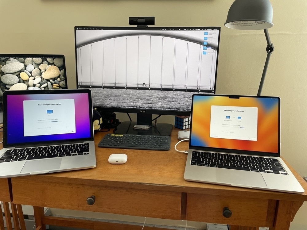 On a wood desk, two laptops have screens open to the Mac Transfer Assistant. Behind them is a large monitor and another laptop, with various desk accessories strewn between. 