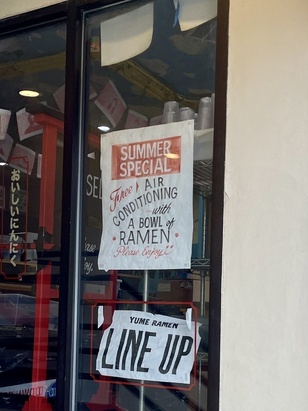A sign in the window of a ramen shop advertises in stylized script, “Summer Special: Free Air Conditioning with a bowl of raman. Please enjoy!”