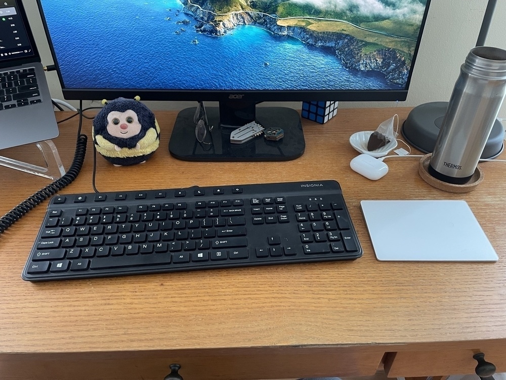 On a wooden desk, a large black wired keyboard with a number pad sits next to a white trackpad. Behind both, a laptop on a stand, a monitor with a mountain and sea desktop image, a stuffed bumblebee, a pair of glasses, two fidget toys, an AirPod charging case, a Rubik’s cube and some tea in a thermos sit. 