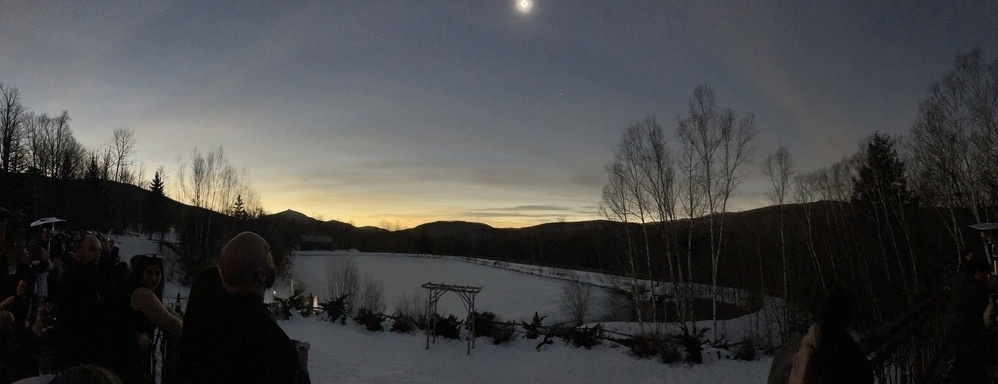 A winter mountain landscape with snow, an open wooden frame, and a frozen lake. The sky is dark; a bright light is cut off in the top center, being the eclipse. 