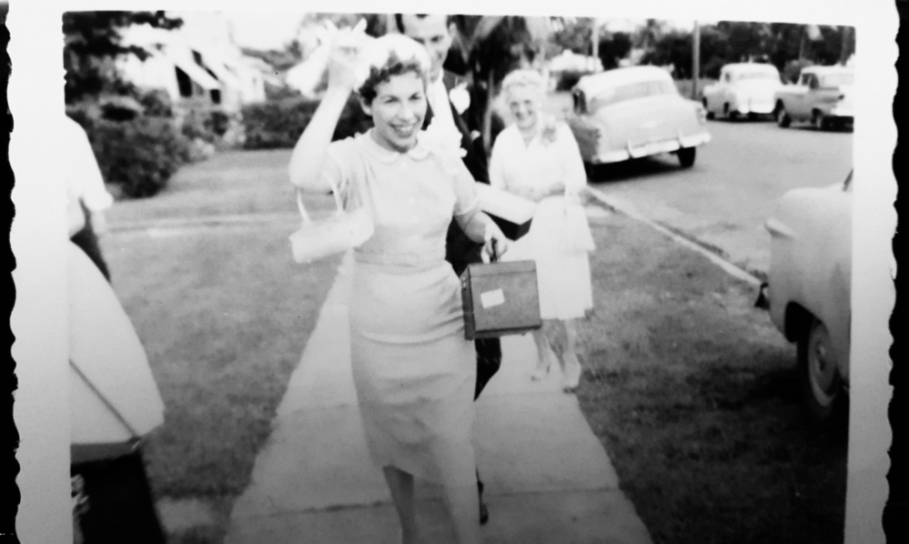 Black and white photo of woman in a light suit and hat, holding a travel bag, man behind here in a dark suit with a buttoniere.