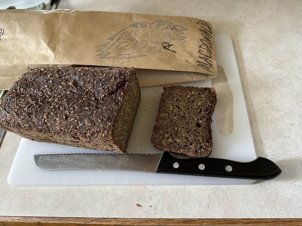 Dark whole grain rectangular loaf of bread, brown paper bag with my name, bread knife 