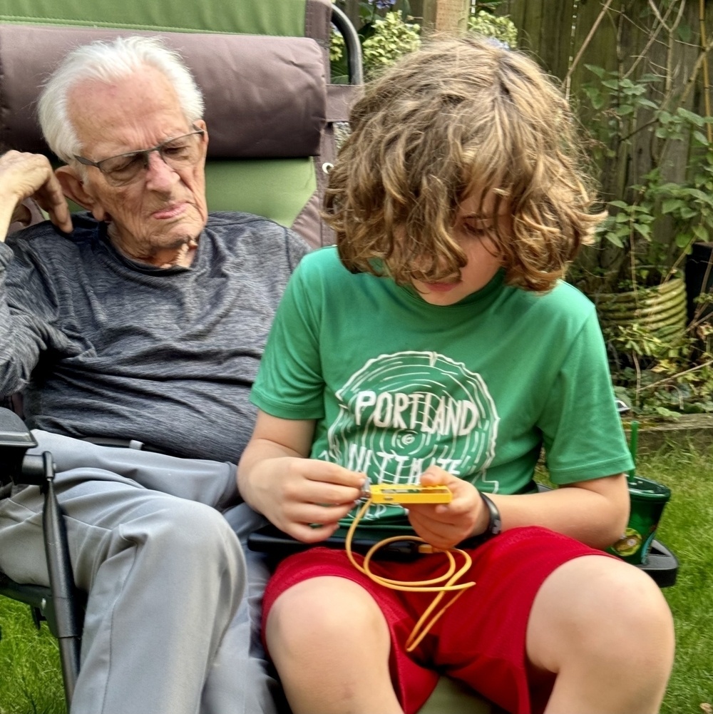 Boy with long hair playing a bright yellow handheld game while elderly man watches. 