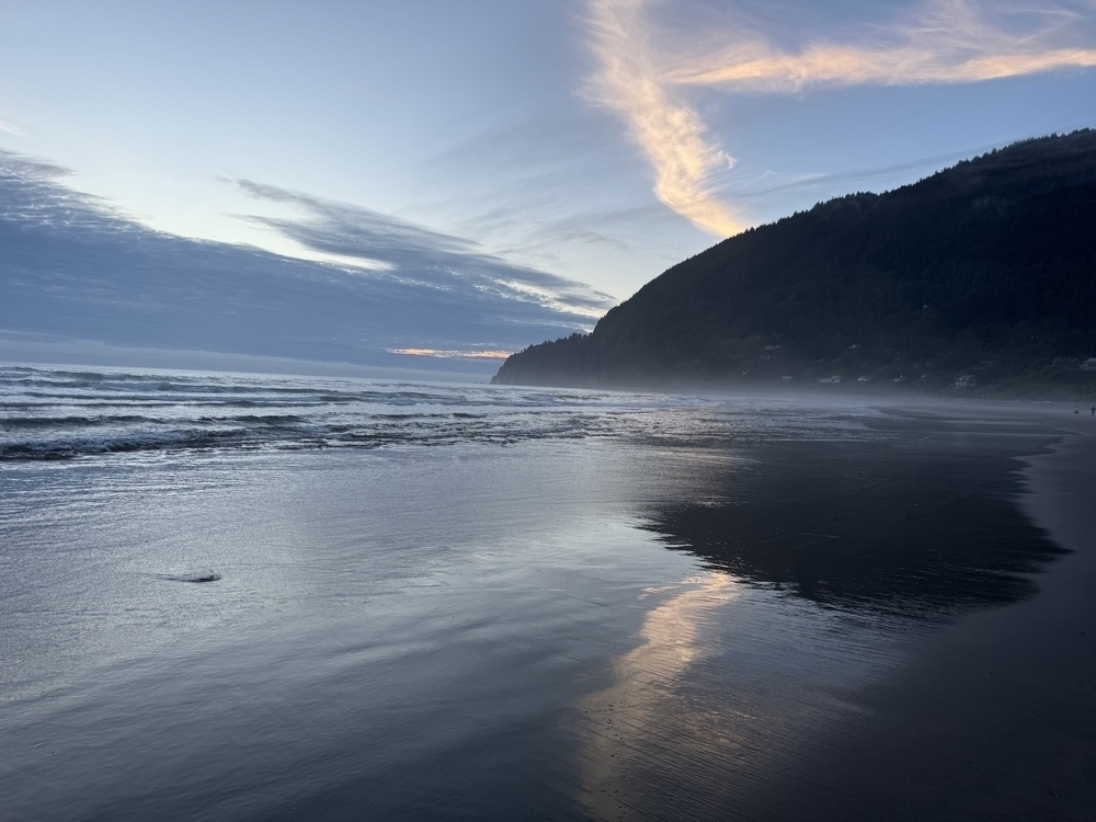 A streak of pink in a dark blue sky, clouds obscuring the sun, all reflected on the wet sands. 