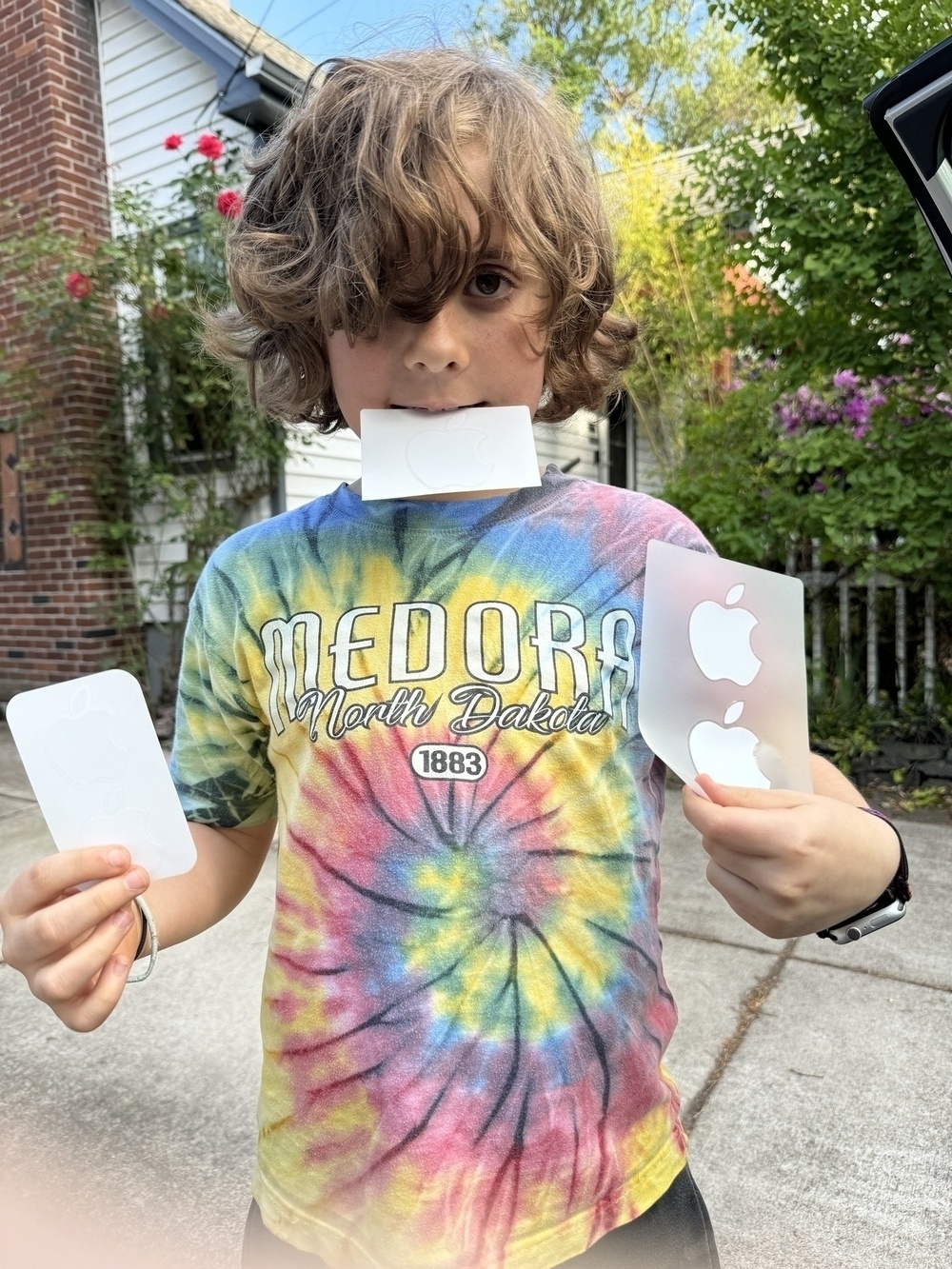 Boy wearing tie-dye tshirt with curly hair in his eyes holding stickers in both hands and his mouth.