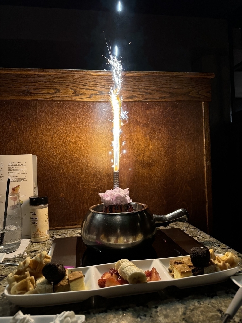 A fondue pot full of melted chocolate. A raspberry cake is in the middle of the chocolate, and a small firework fountain mounted on top of the cake is shooting out sparks.