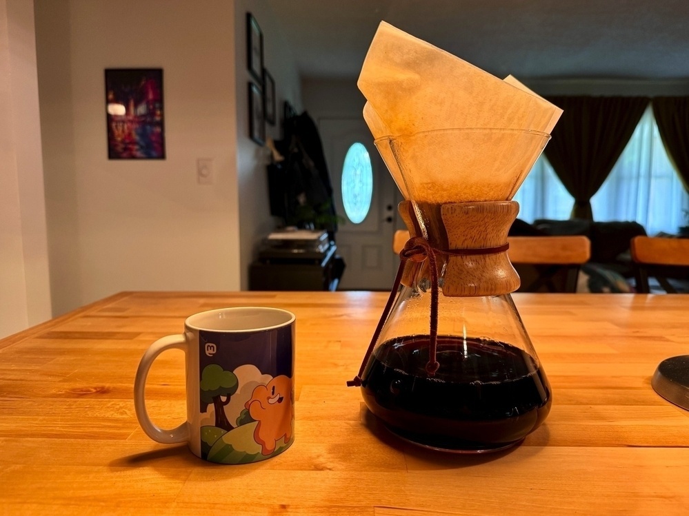 A Mastodon themed mug next to a half-full Chemex branded carafe still brewing coffee. The Chemex has a paper filter funneled over the top, dripping coffee into the carafe below. 