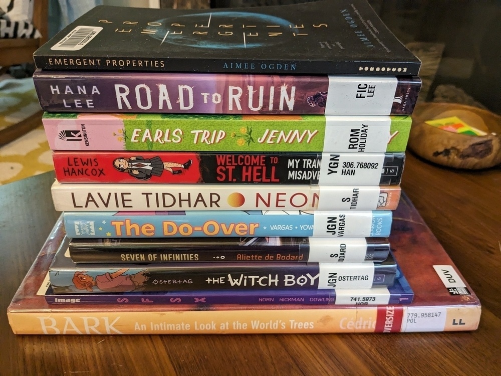 stack of library books including 3 novels, 2 novellas, and 4 graphic novels, mostly queer for Pride Month
