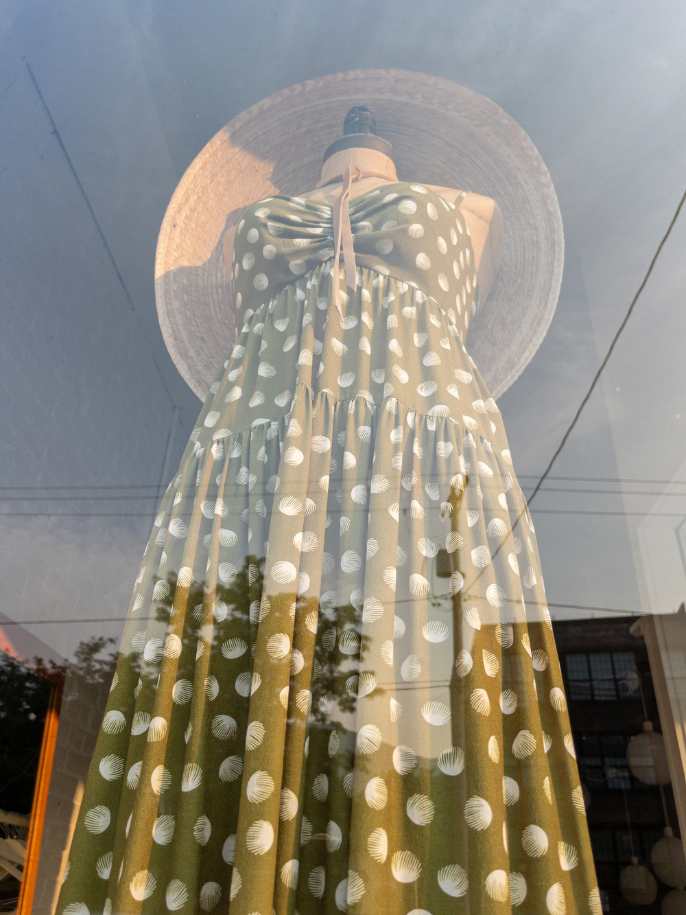 Woman’s sundress, green/brown with beige circular graphic flowers, gathered at the hips and again at the bust, with broad brimmed woven hat on a mannequin. Shot looking up the length of the dress with sky and building reflection on the shop window overlaid.