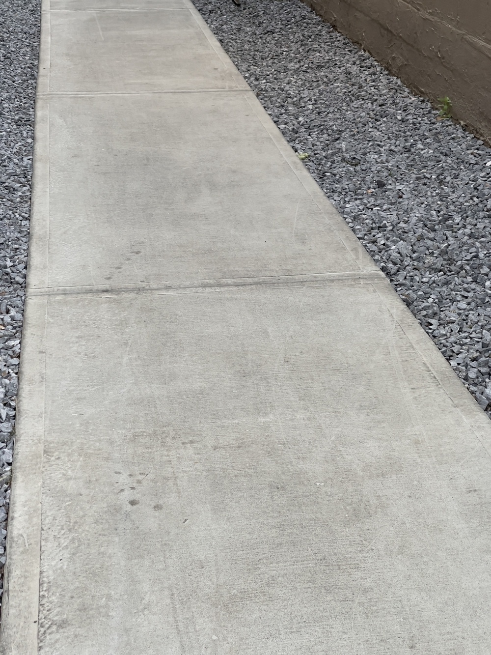 Sidewalk with gravel on both sides and part of a brick wall. Parallel lines converge towards the left corner.