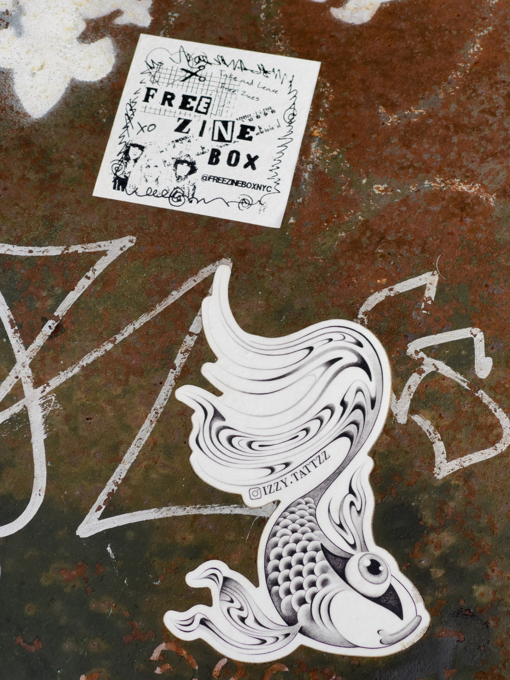 Stickers on a metal surface. The top is for “Free Zine Box.” The bottom is an oriental stylized drawing of a Koi fish.