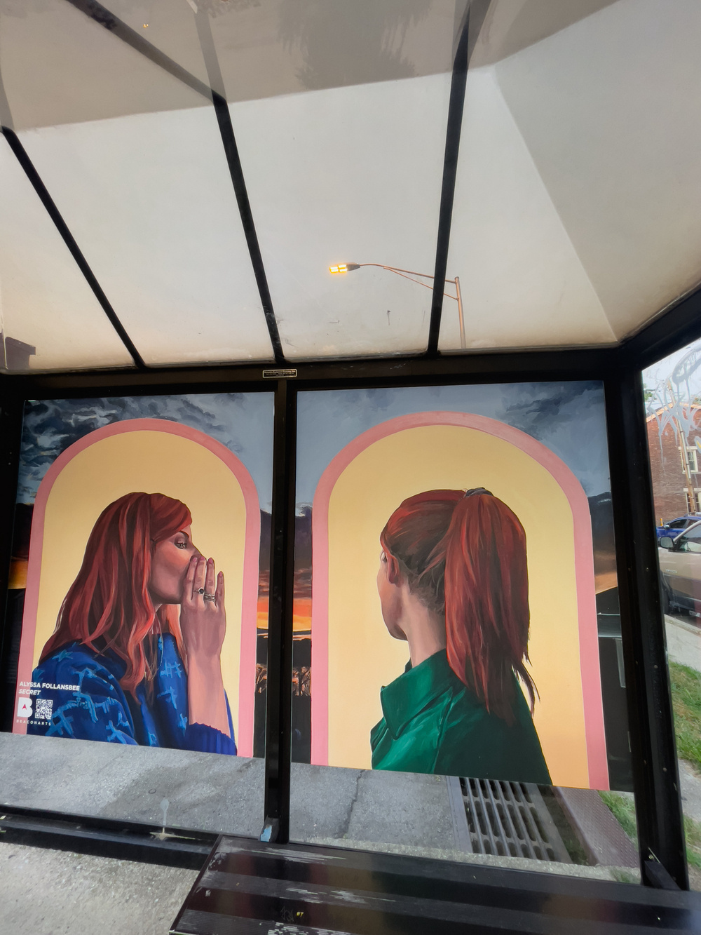 Bus shelter with artwork. Paintings of two women, the woman on left appears to be whispering into the ear of the woman on the right. The transparent plexi roof with streetlight in the distance.