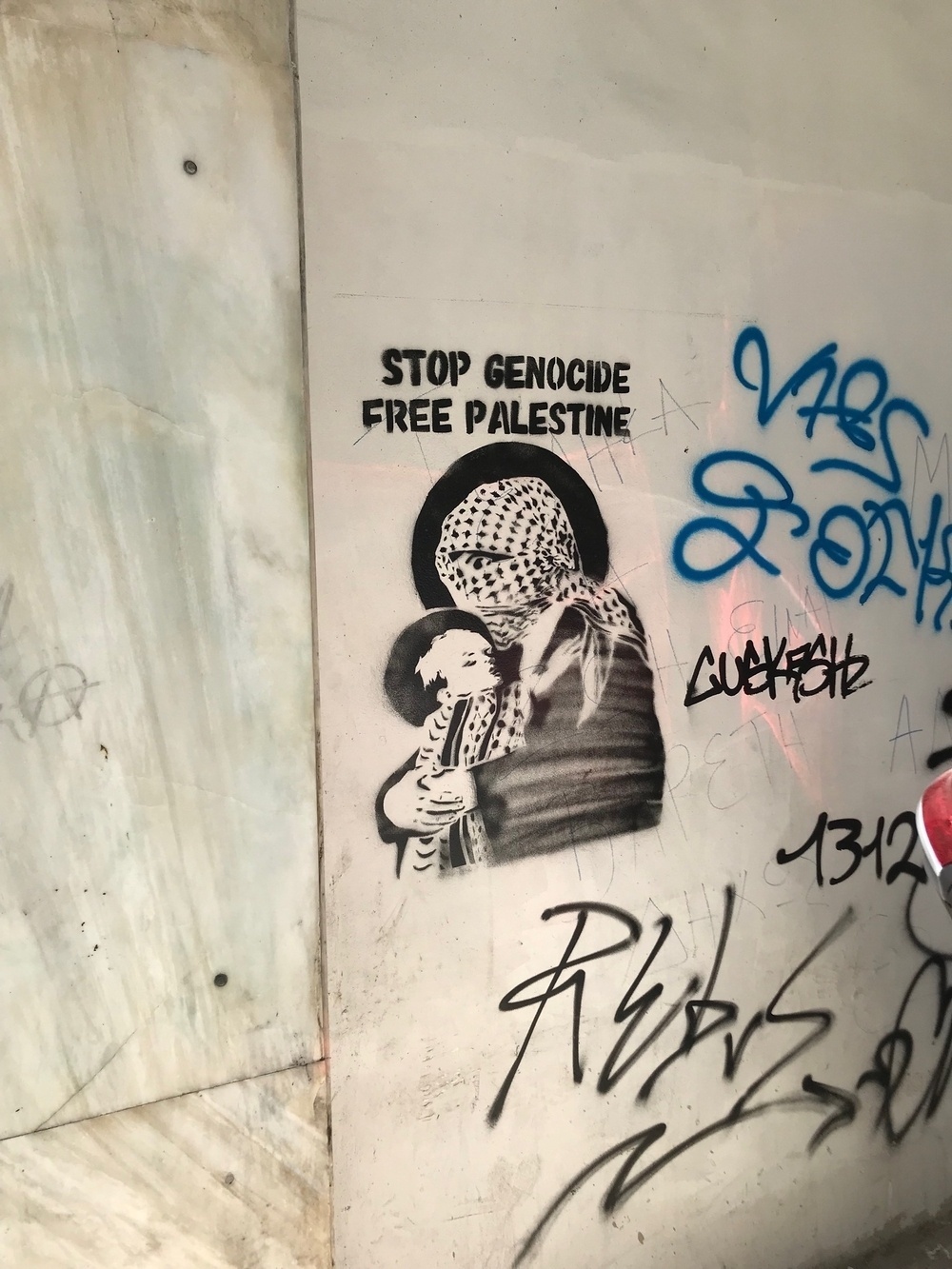 Graffiti in Athens with text reading "Stop Genocide, Free Palestine" above a stencil drawing of a person with their head and face completely wrapped in a keffiyeh, holding a child is wrapped in a keffiyeh. Both of their heads are haloed by a black circle