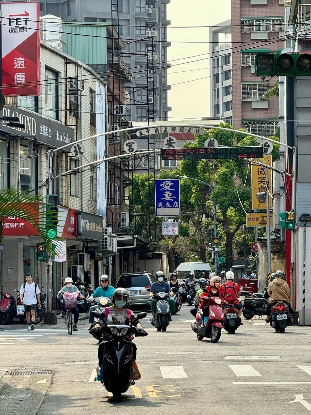 A small road junction, with several motorcycles coming towards the camera, and a blue sign hanging in the distance reading 愛妻洗衣店