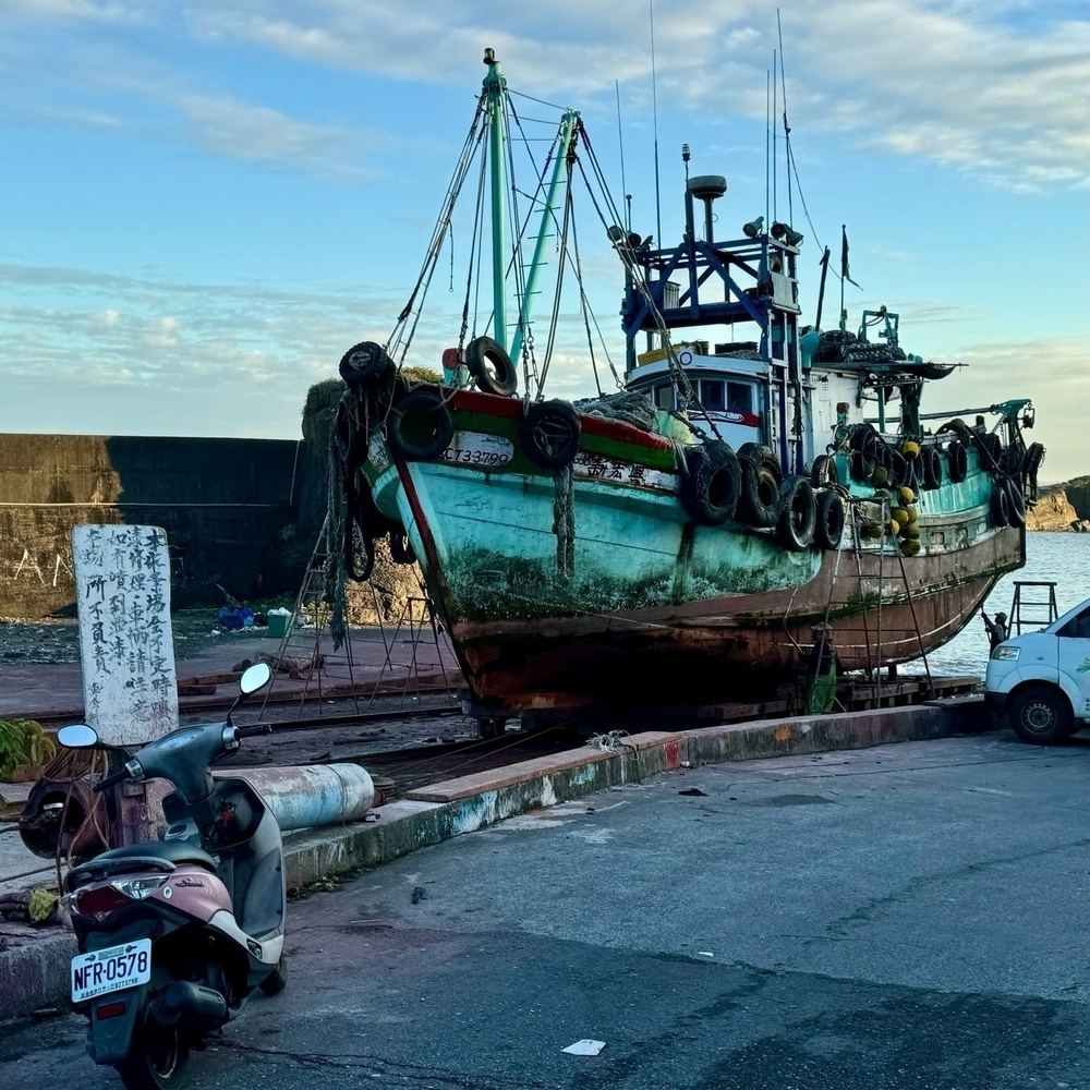 A green fishing boat up on the dry dock being repaired/painted, beside a white sign relinquishing responsibility for paint that gets splashed on any cars parked nearby.