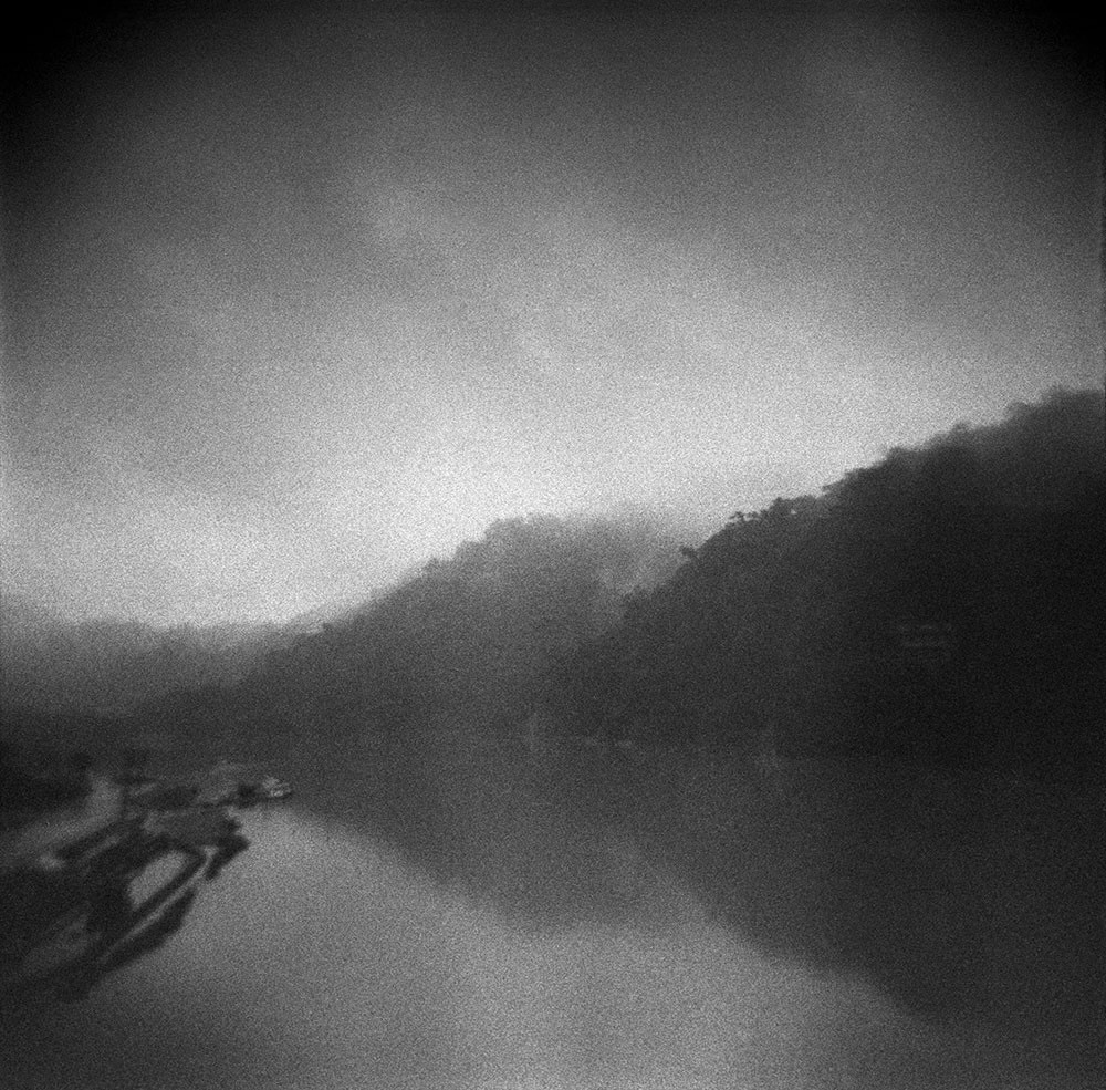 Black and white misty photo taken from a bridge, of a river, bounded by mountains on the right side.