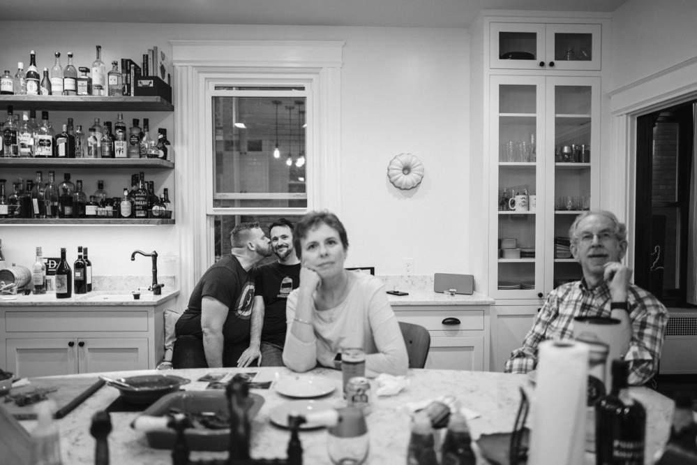 A man smootches his husband in the background of a kitchen while his parents look at the camera seated at the island. They're out of focus. - Leica Q