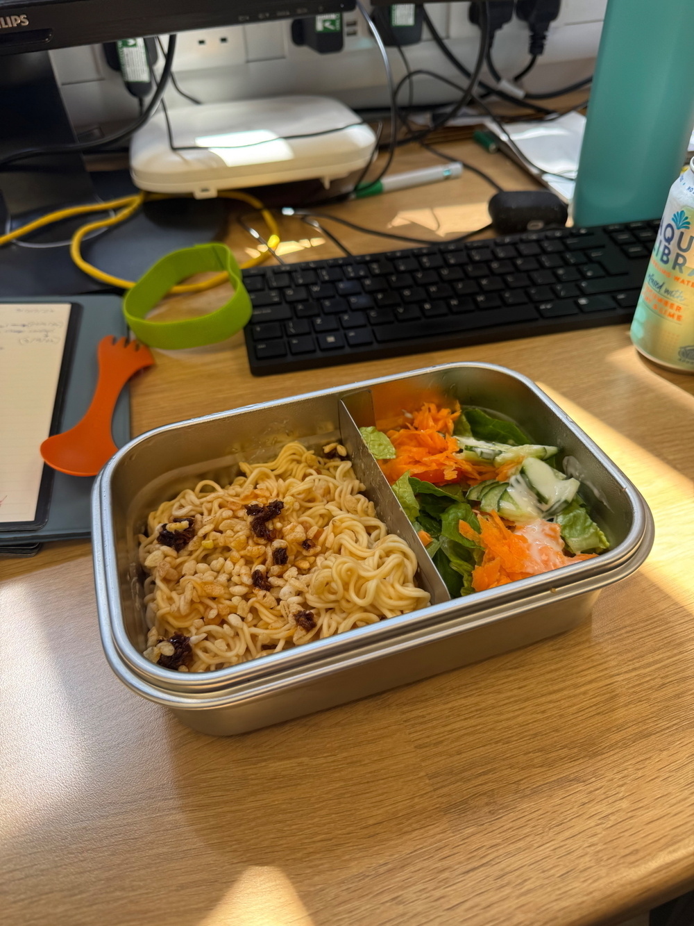 A divided metal lunch container with peanut noodles topped with chilli flakes on one side, and a salad with lettuce, cucumber, shredded carrots, and dressing on the other side.