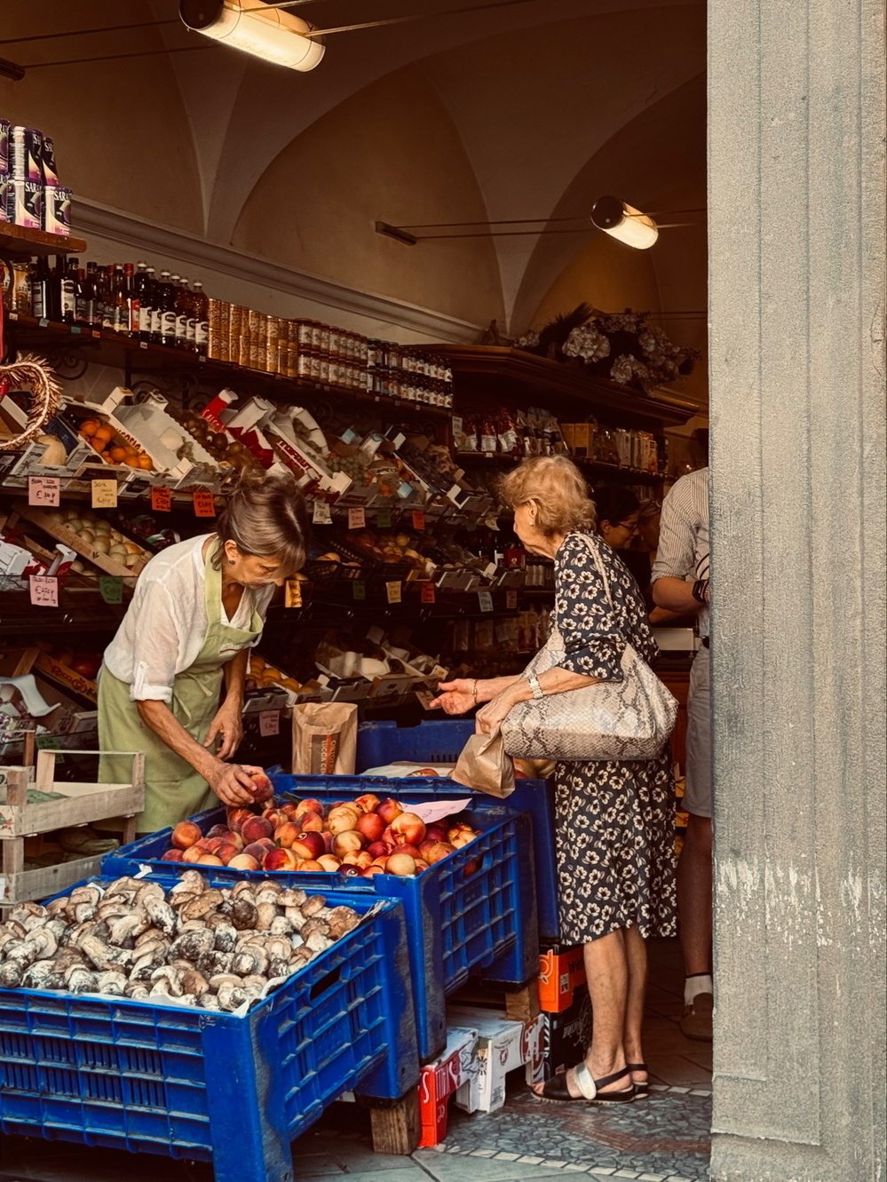 Two women are interacting inside a market or small grocery store. One woman, wearing an apron, is helping the other woman, who is carrying a snakeskin-patterned bag, with selecting produce.