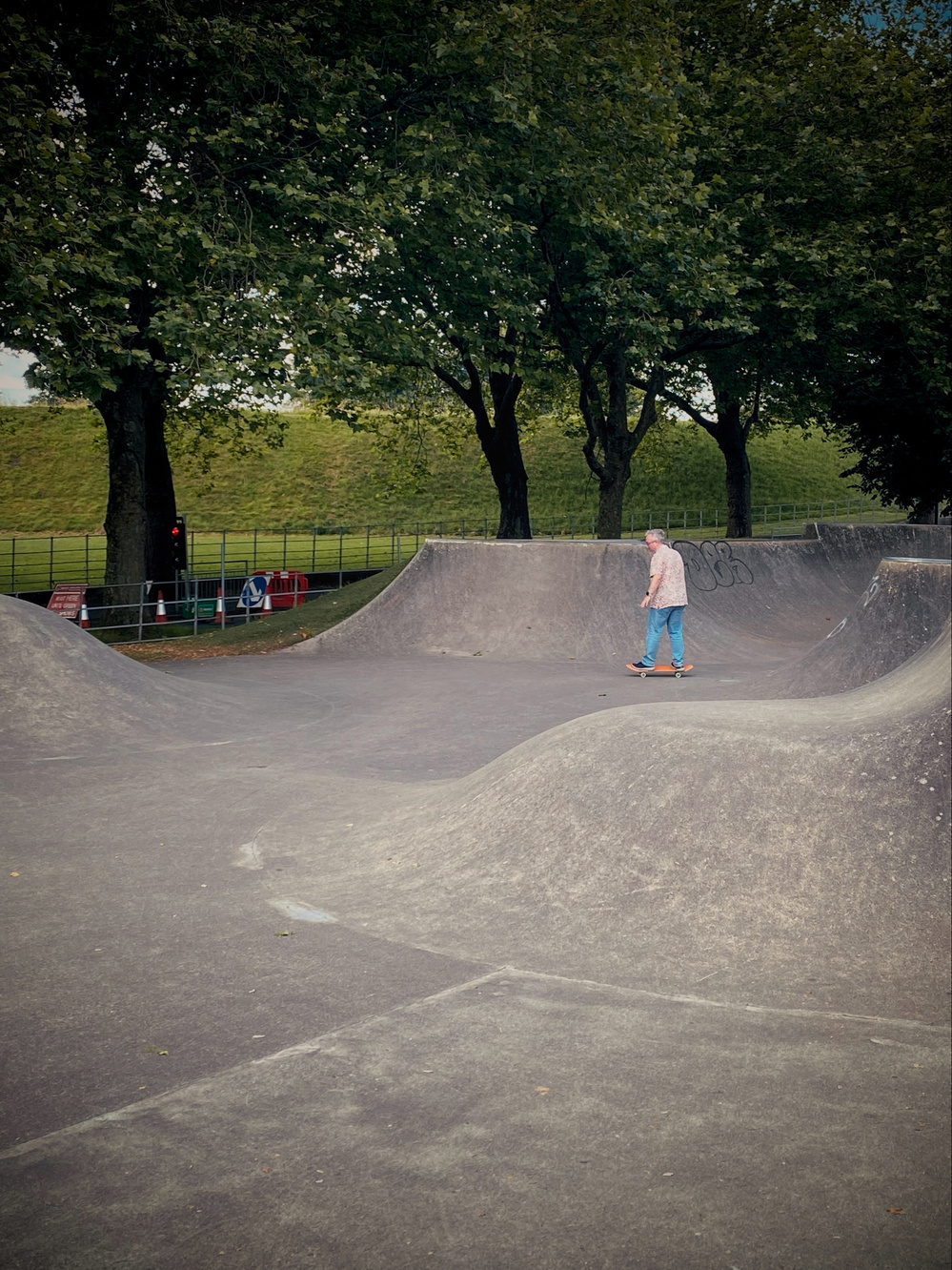 An individual is skateboarding on a concrete skatepark structure surrounded by green trees. There is a grassy hill in the background, and safety barriers with traffic cones are visible to the left. The sky is overcast, and the environment appears calm and serene