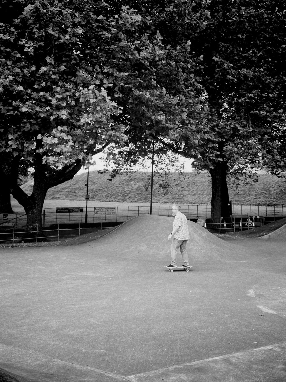 A black-and-white image of an older man skateboarding on a concrete ramp in a skatepark. The backdrop includes large, leafy trees and some fencing, with signs visible in the background.