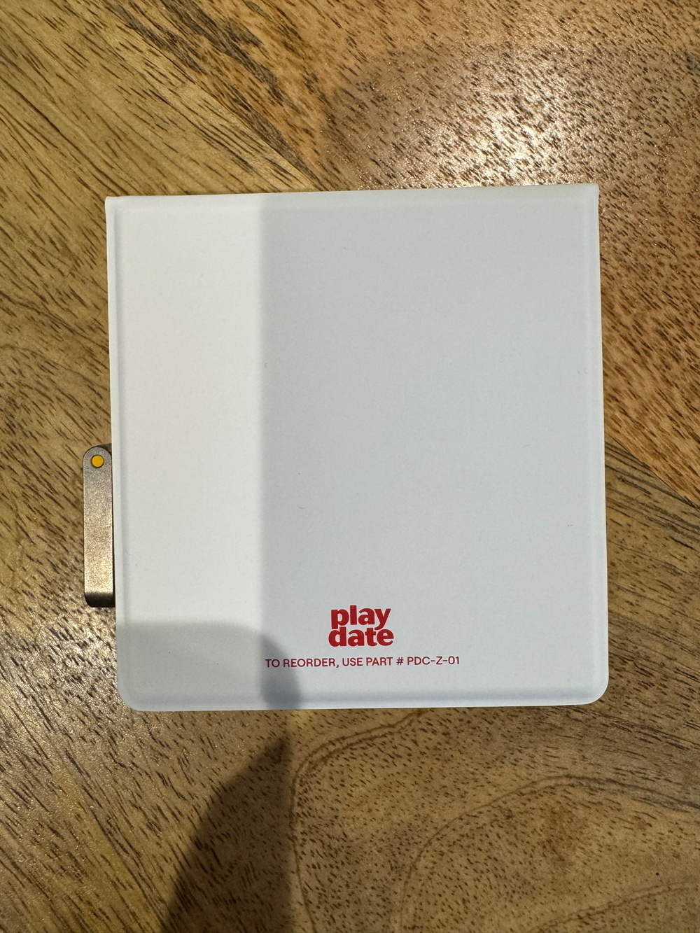 A white object with a partial metal hinge on the left side, featuring red text that reads “play date” and “TO REORDER, USE PART # PDC-Z-01” on a wooden surface.