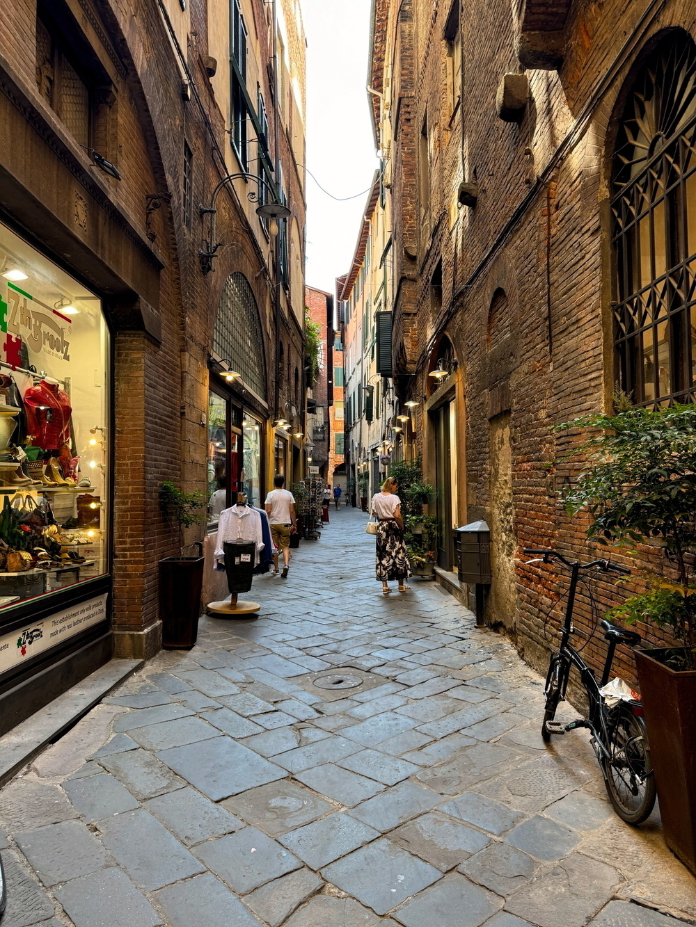 A narrow cobblestone alleyway flanked by tall, historic brick buildings. The alley features small shops with window displays, a parked bicycle, and a few pedestrians walking through it. The image captures a quaint European street scene