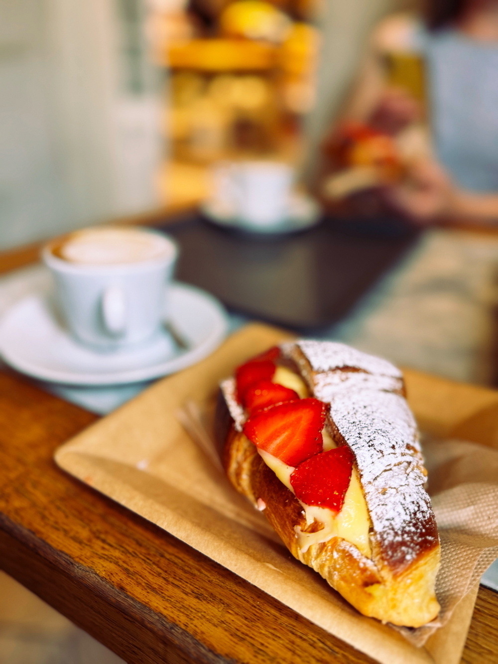 Close-up of a custard-filled pastry topped with fresh strawberries and powdered sugar on a wooden table, accompanied by a cup of coffee in the background.