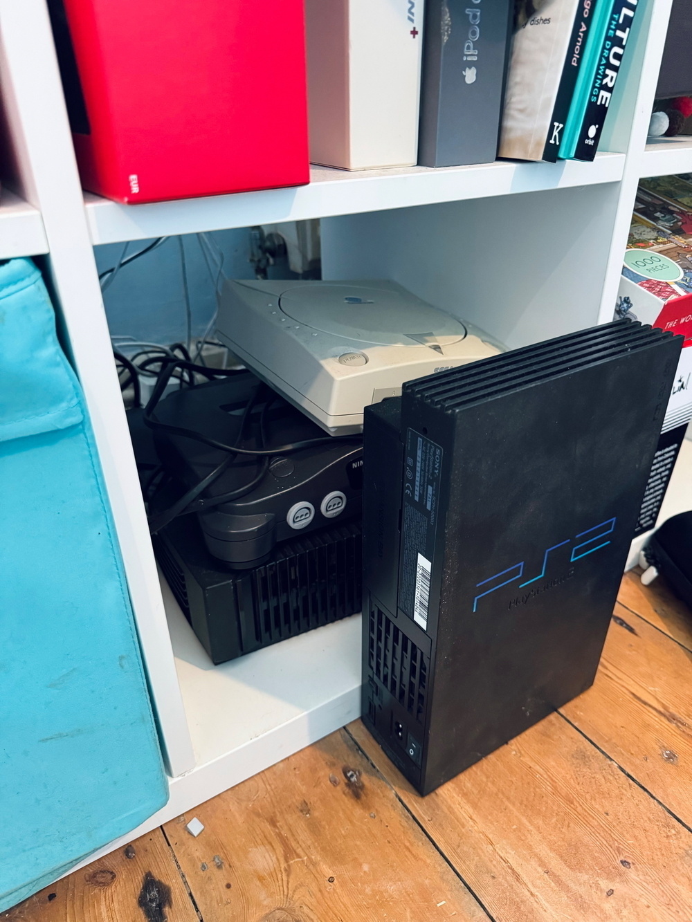 A shelf holding several vintage gaming consoles, including a Sony PlayStation, a Nintendo 64, and a PlayStation 2. Books and boxes, including an iPod box and one labeled “Eva,” are also visible.