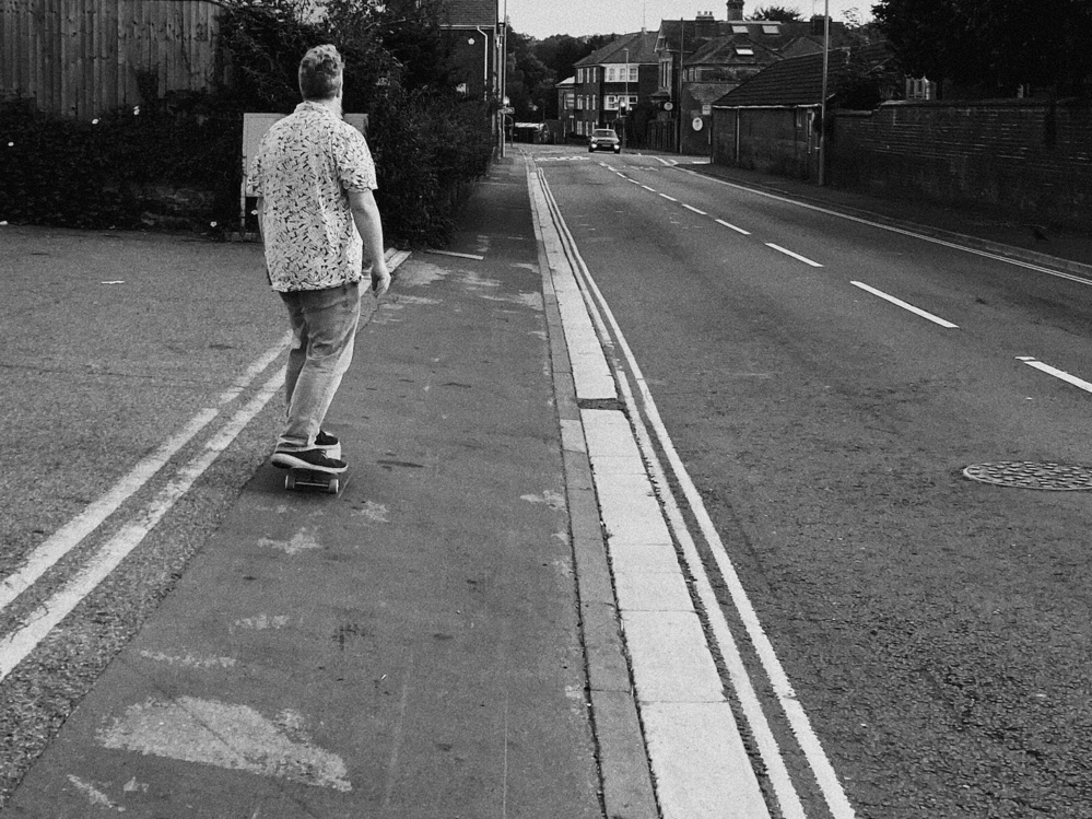 A black and white photograph of a person skateboarding on a sidewalk beside a street. The person is wearing a patterned short-sleeve shirt and jeans, and they are seen from behind. There is a road with a parked car in the distance,