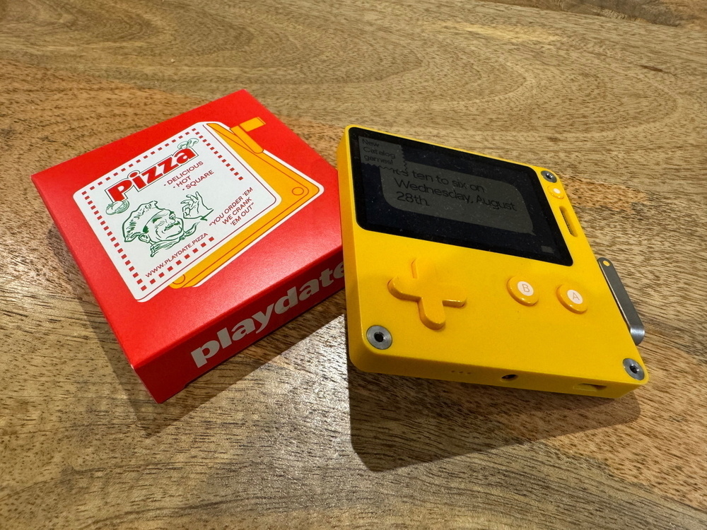 A Playdate handheld gaming console in yellow color is placed next to a red box labeled “Pizza” with a cartoon chef and the Playdate logo.