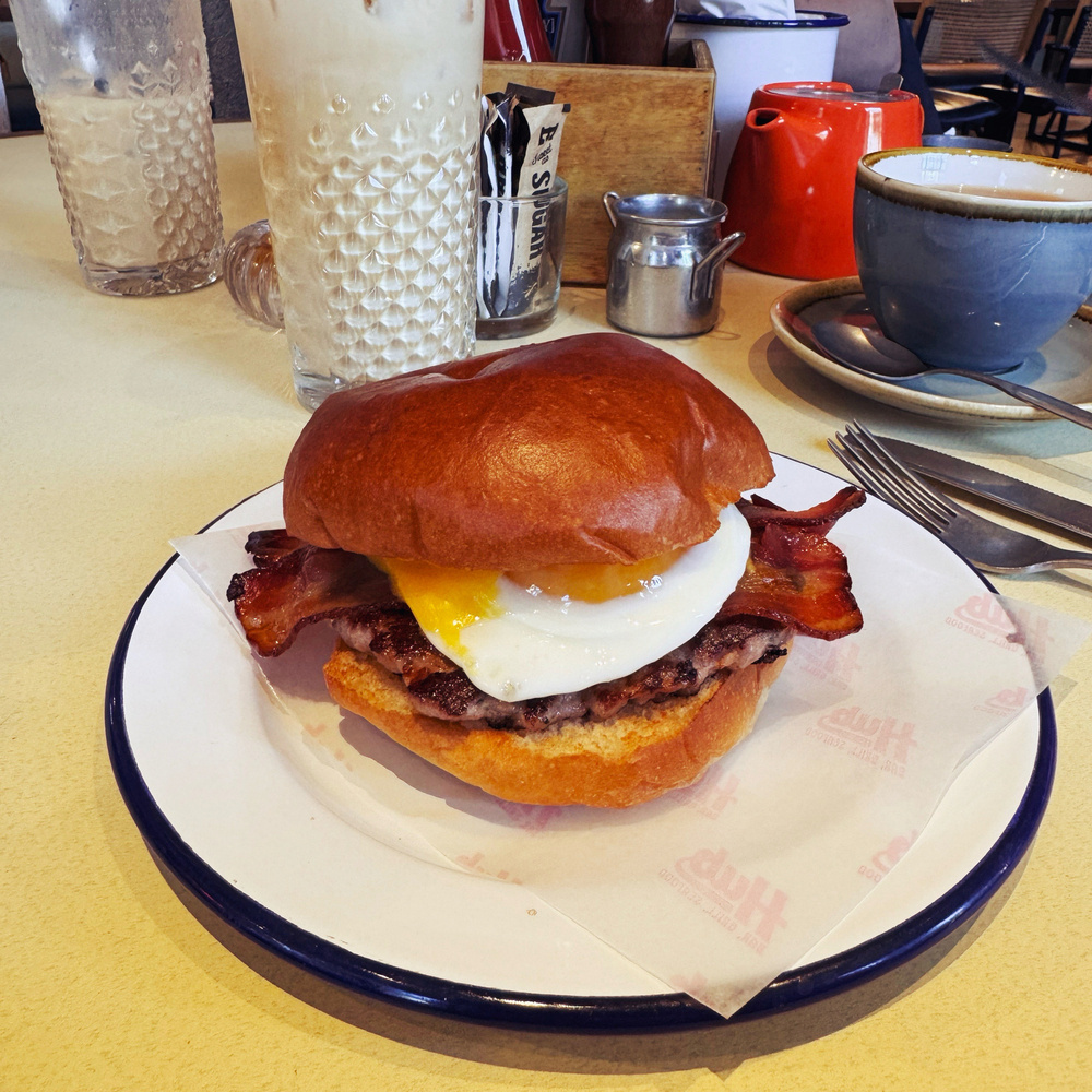 A close-up image of a breakfast burger featuring a fried egg, bacon, and a beef patty, served on a brioche bun. In the background, there are glasses, a teapot & coffee cup on a saucer.