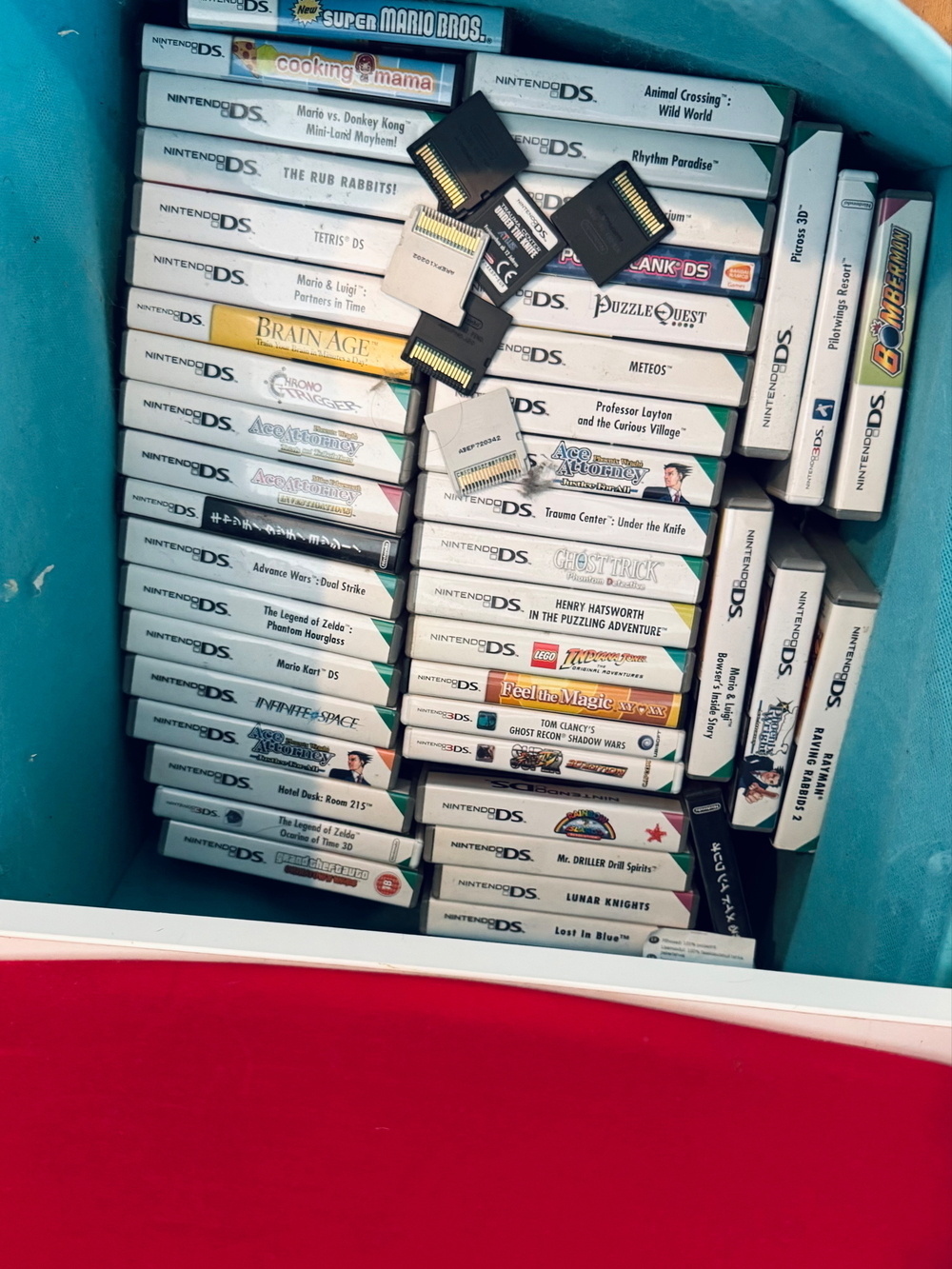 A collection of Nintendo DS and 3DS game cases organized in a box. Several game cartridges are stacked on top of the cases. The games included are titles like Super Mario Bros., Cooking Mama, Mario Kart DS, Chrono Trigger